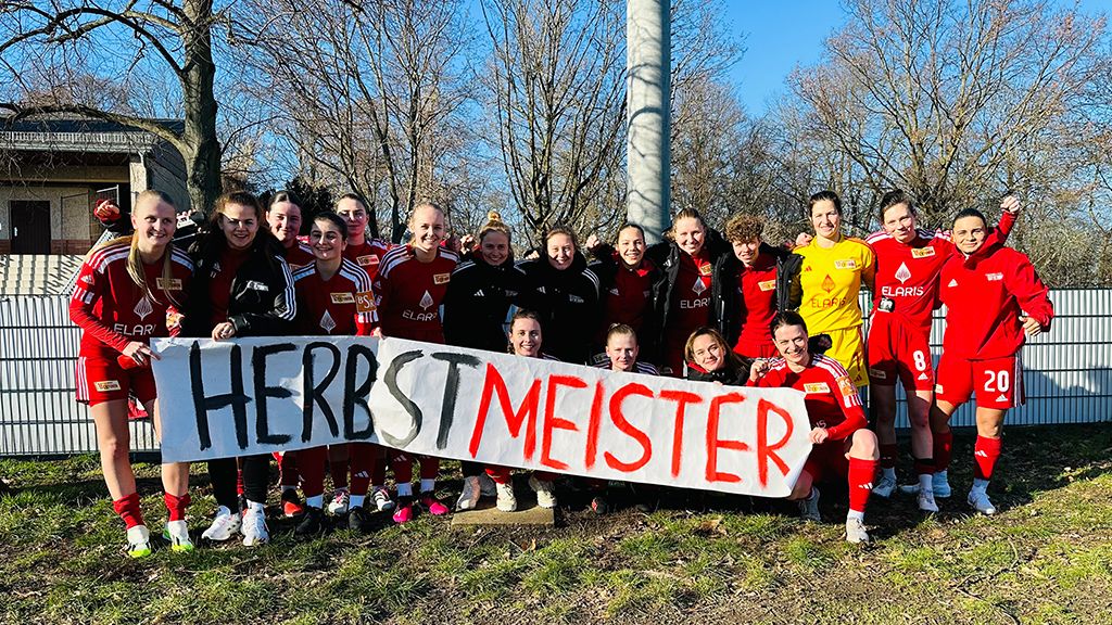 Mannschaft im roten Trikot hält ein Schild mit der Aufschrift "HERBSTMEISTER", umringt von Bäumen und einem Sportplatz.