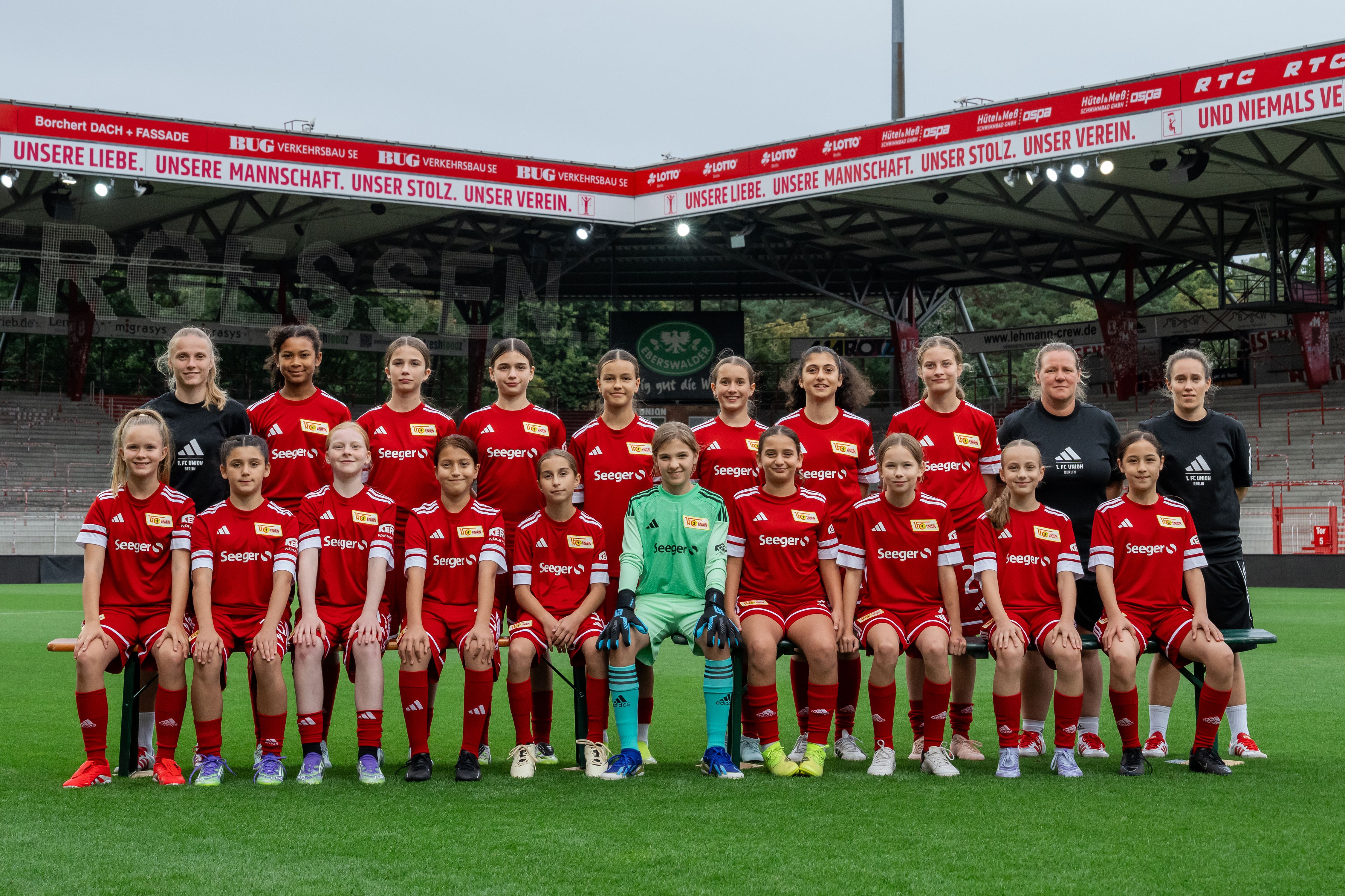 Mannschaftsfoto eines Jugendfußballteams in roten Trikots, auf einem Stadionrasen, mit Trainer und Spielerinnen in zwei Reihen.