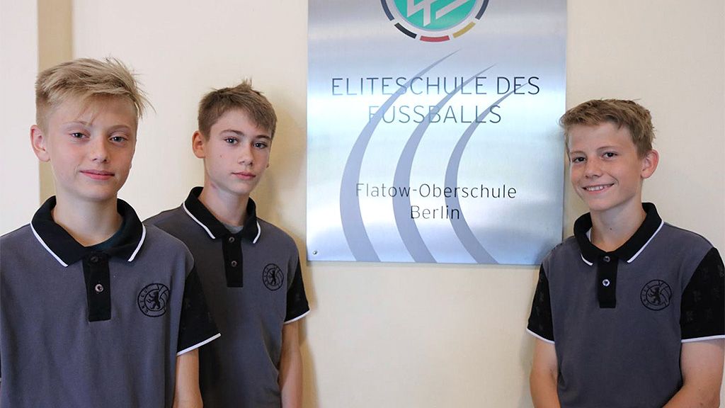 Drei Jungen stehen vor einem Schild mit der Aufschrift "Eliteschule des Fußballs" in Berlin.