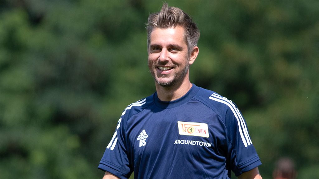 A smiling man in a blue sports jersey with a football club logo is standing outdoors.