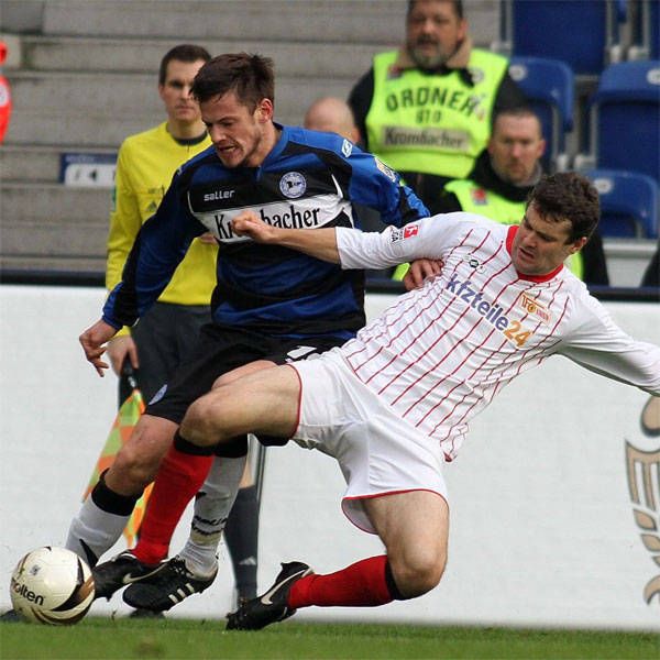 Zwei Fußballspieler im Zweikampf, einer fällt, während der andere ihn festhält; Zuschauer im Hintergrund.