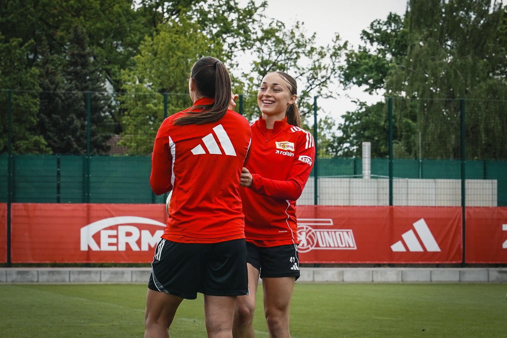 Zwei Sportlerinnen in roten Trikots lachen und interagieren auf einem Fußballfeld mit einem grünen Hintergrund.
