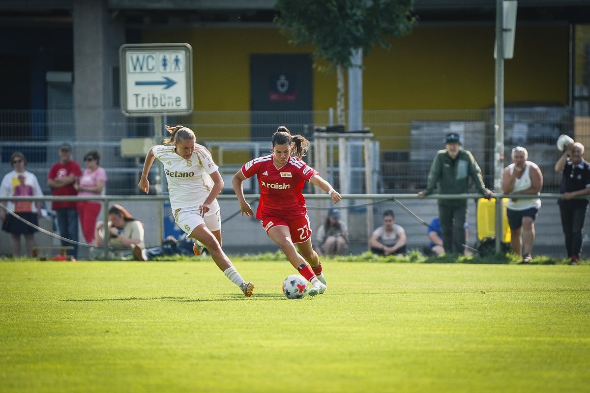 Zwei Fußballspielerinnen verfolgen sich auf einem grünen Spielfeld, während Zuschauer im Hintergrund zuschauen.