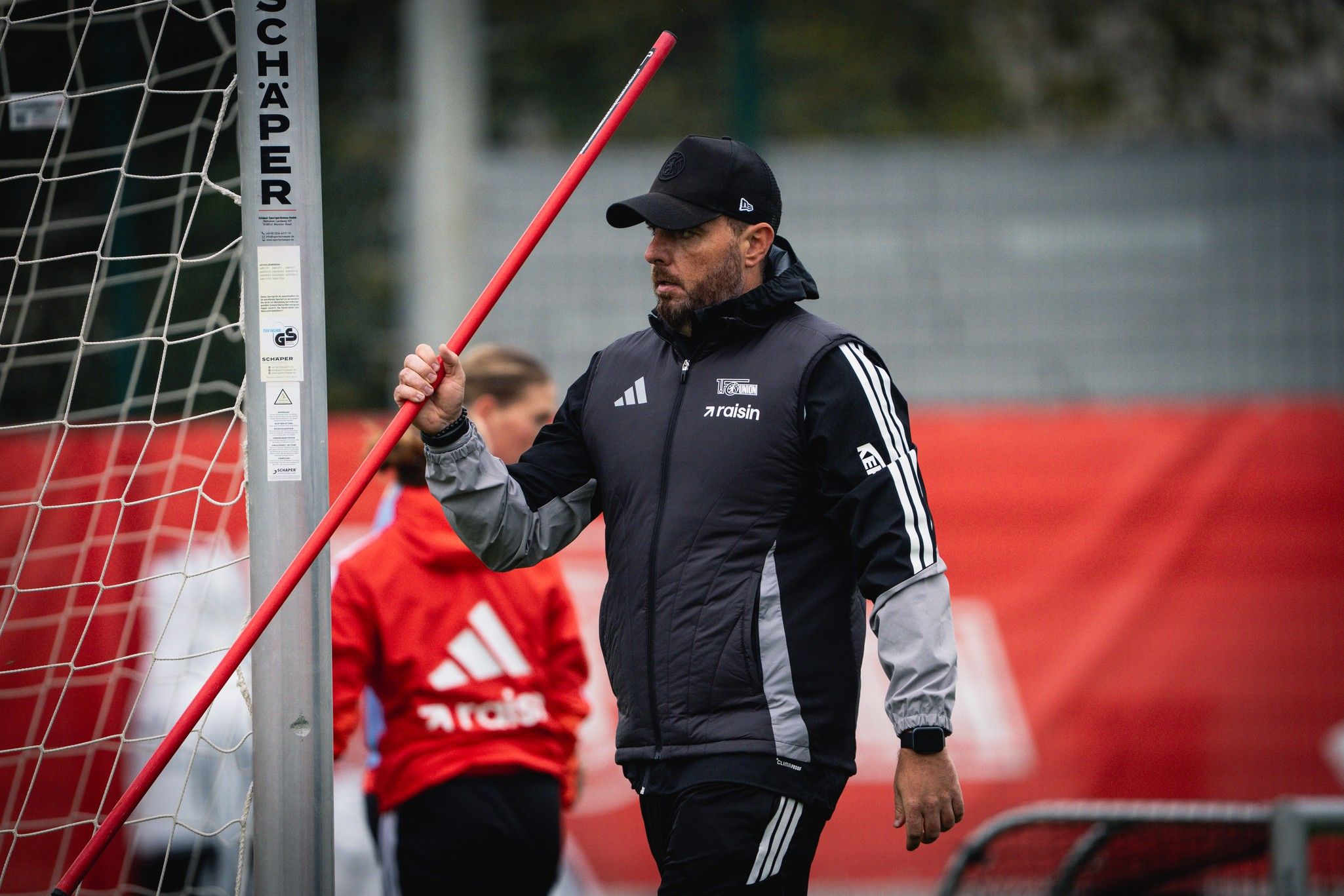 Ein Trainer in Sportkleidung hält einen langen roten Stab und geht an einem Fußballtor vorbei. Im Hintergrund sind Spielerinnen zu sehen.