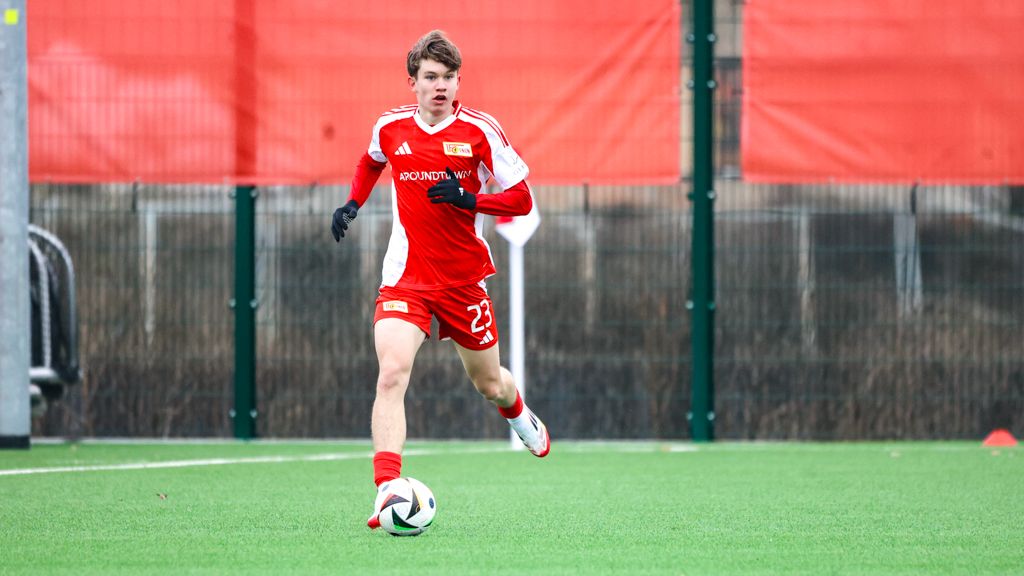 Ein junger Fußballspieler in rotem Trikot sprintet über ein Kunstrasenfeld.