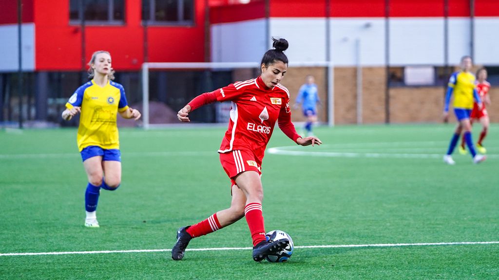 Eine Spielerin trägt rote Kleidung. Sie dribbelt den Ball auf dem Fußballfeld. Eine Gegnerin in blauer und gelber Kleidung folgt ihr.