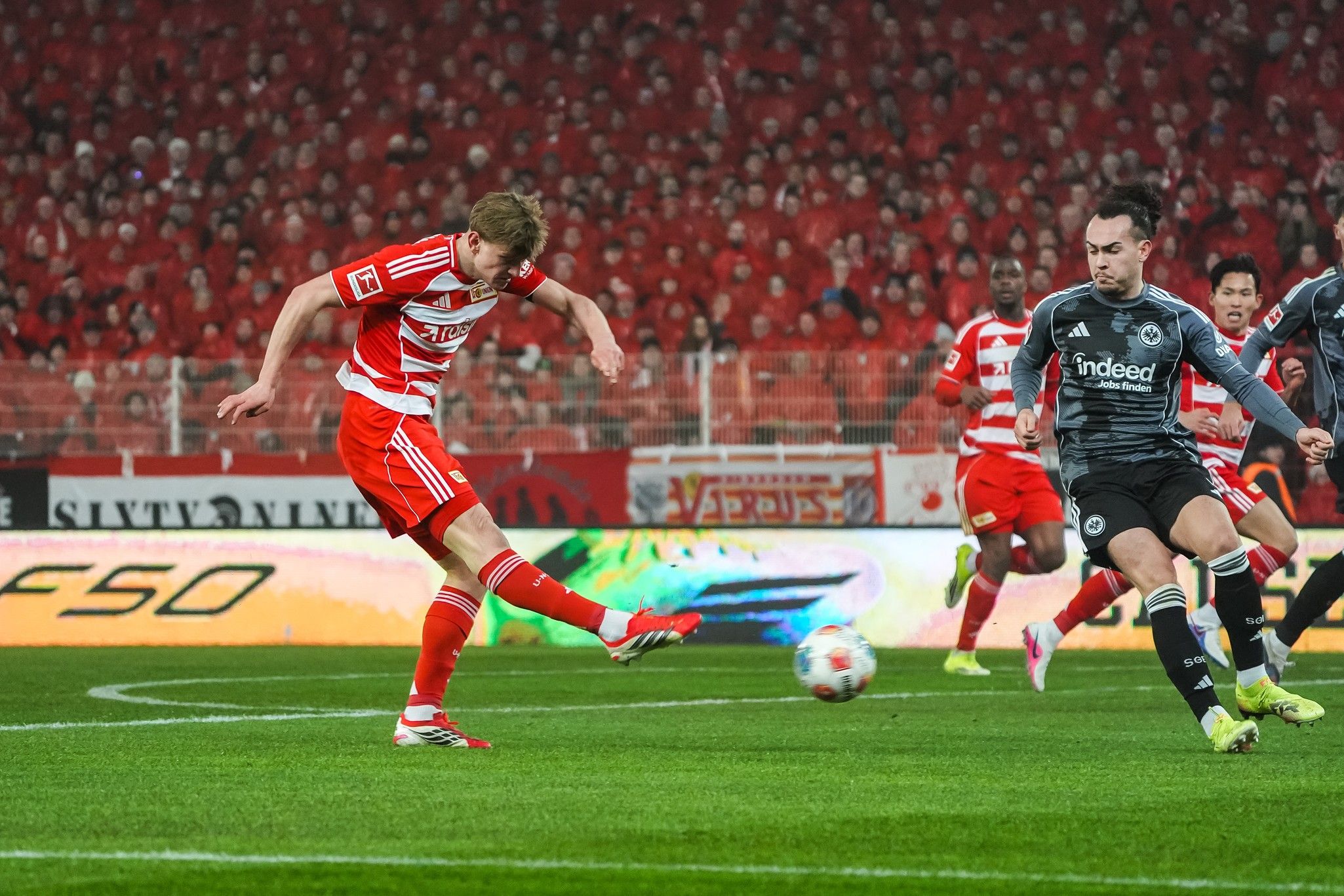 Spielszene mit einem Spieler in roter und weißer Fußballuniform, der auf das Tor schießt, während ein gegnerischer Torwart vorbereitet ist.