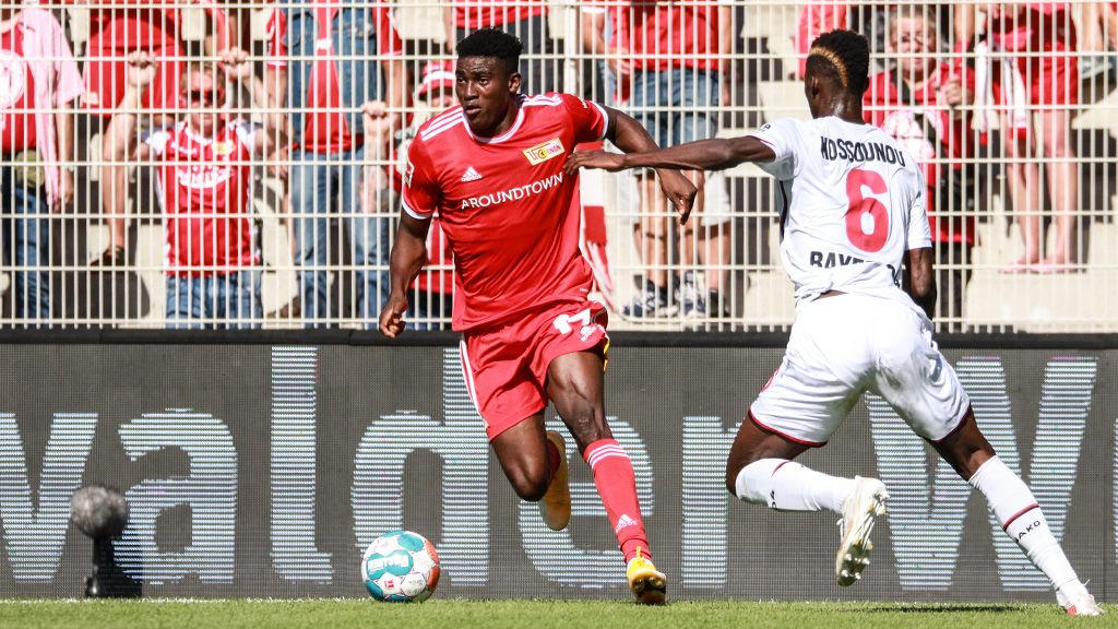 Ein Fußballspieler trägt ein rotes Trikot. Er rennt mit dem Ball. Ein Spieler in weißem Trikot läuft hinter ihm her.