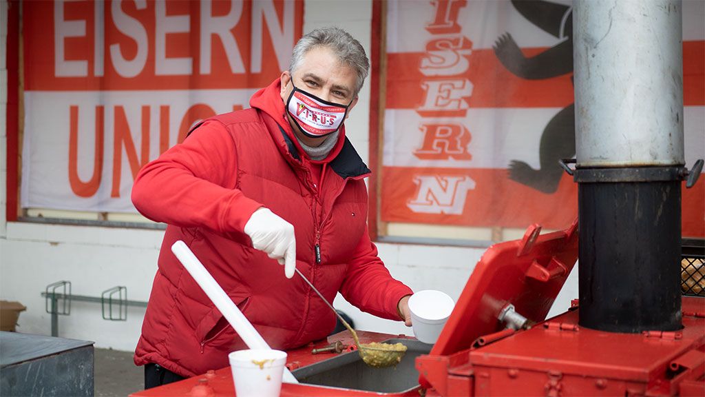 Mann in roter Jacke und Maske serviert Essen aus einem mobilen Verkaufsstand. Hintergrund mit Schriftzug „EISERN UNION“.