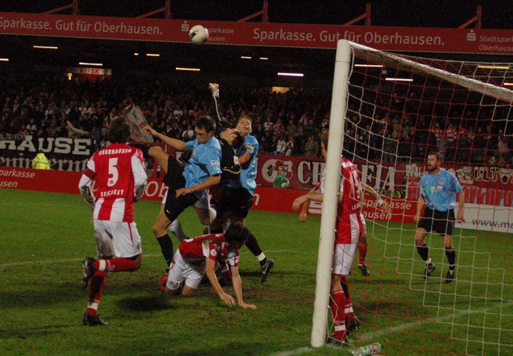 Spielszene im Fußballstadion: Ein Torwart springt nach dem Ball, während Spieler auf dem Platz um ihn herum kämpfend agieren.