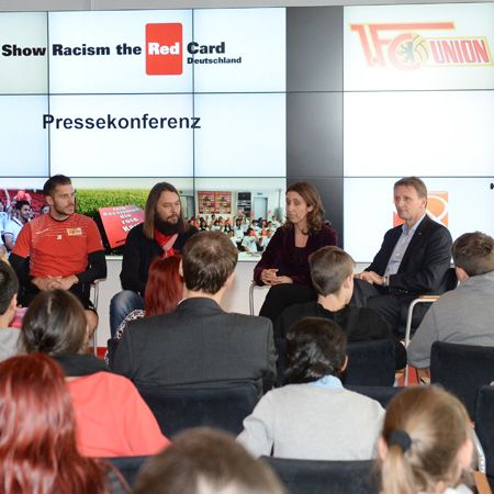 Pressekonferenz mit mehreren Personen, im Hintergrund ein Bildschirm mit dem Slogan "Show Racism the Red Card Deutschland".