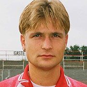 Porträt eines jungen Mannes mit blonden Haaren, in einem roten Sporttrikot, mit neutralem Gesichtsausdruck auf einem Sportgelände.