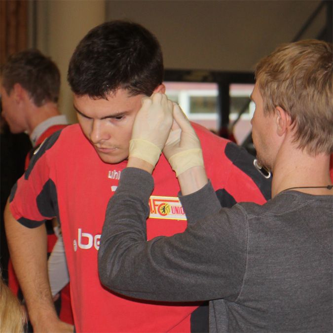 Ein junger Mann in einem roten Trikot erhält eine Behandlung am Ohr von einer Person in Handschuhen.