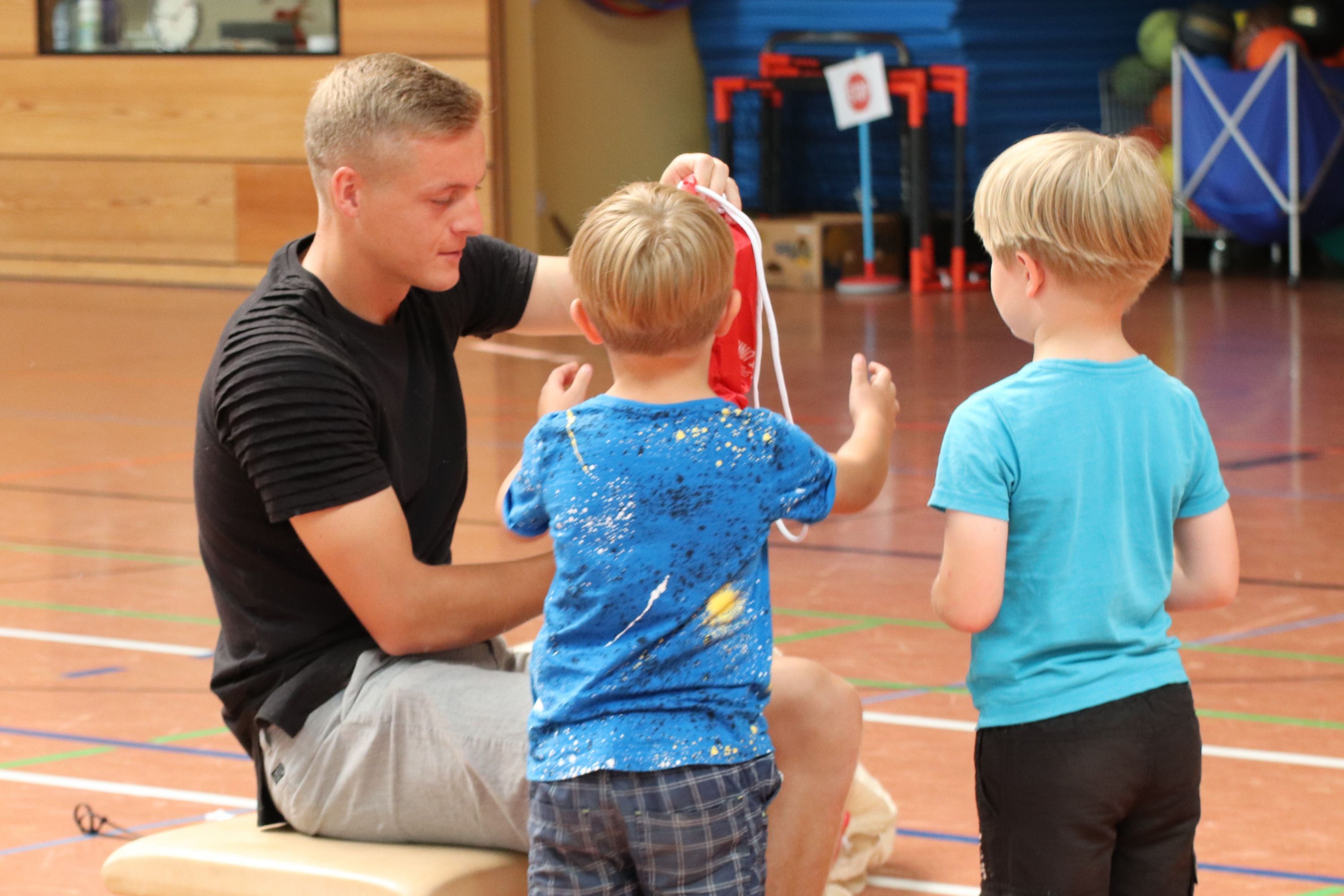 Ein Erwachsener sitzt auf einer Bank und zeigt zwei Kindern einen roten Sack in einer Sporthalle.