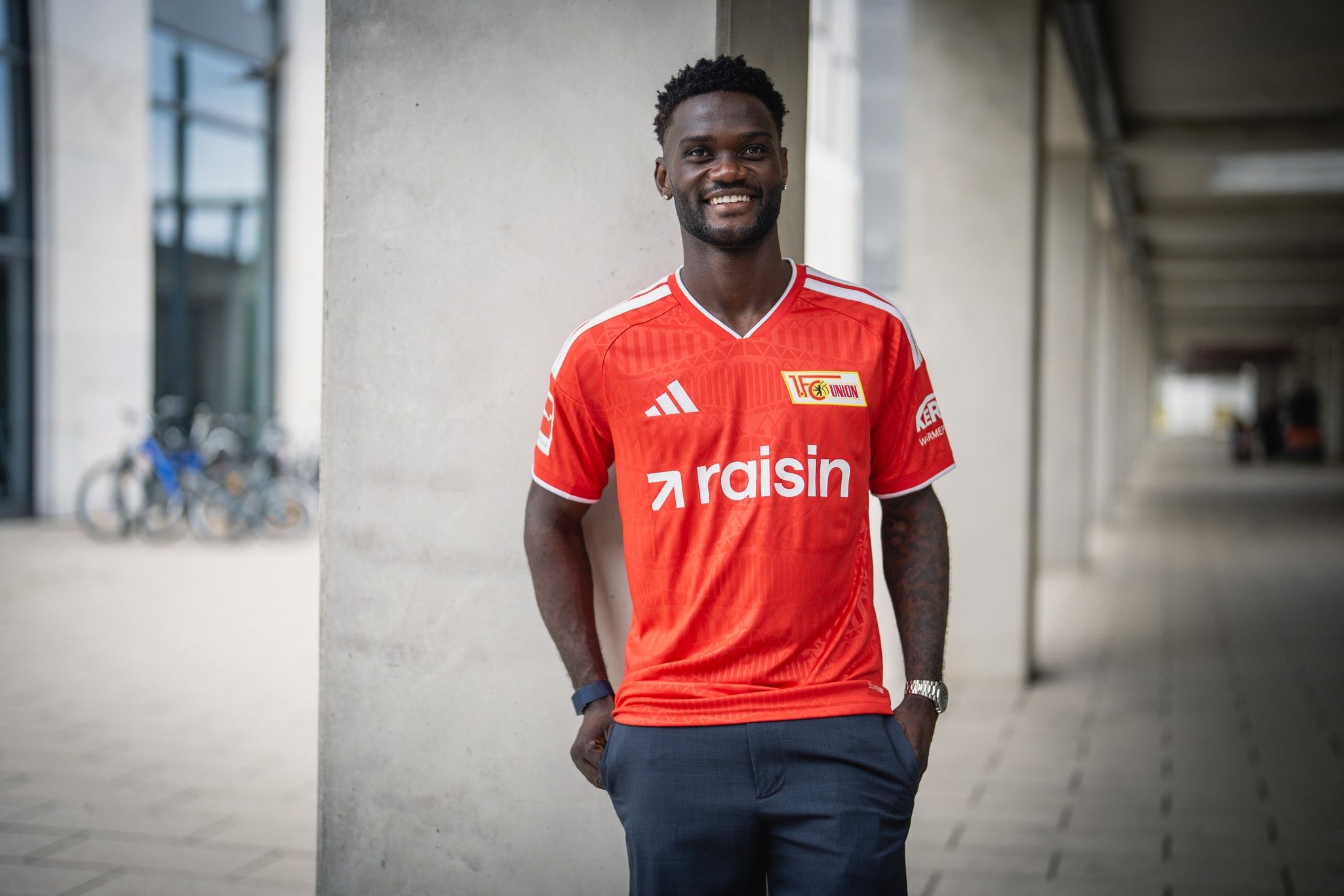 Mann in rotem Fußballtrikot mit "raisin"-Logo steht lächelnd an einem Betonsäulen-Treppenaufgang.