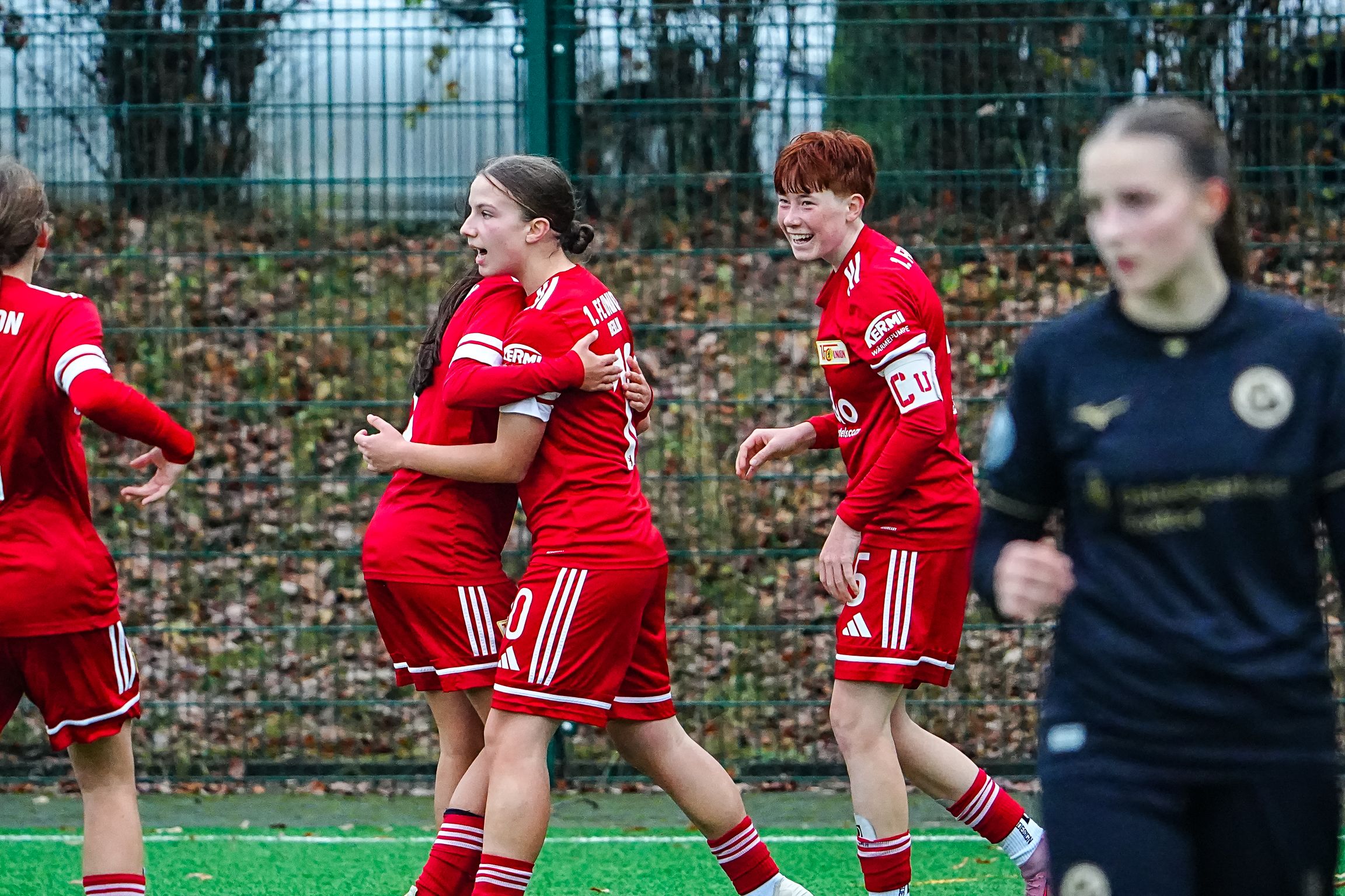 Zwei Fußballspielerinnen in roten Trikots umarmen sich. Eine andere Spielerin freut sich. Im Hintergrund ist eine vierte Spielerin.