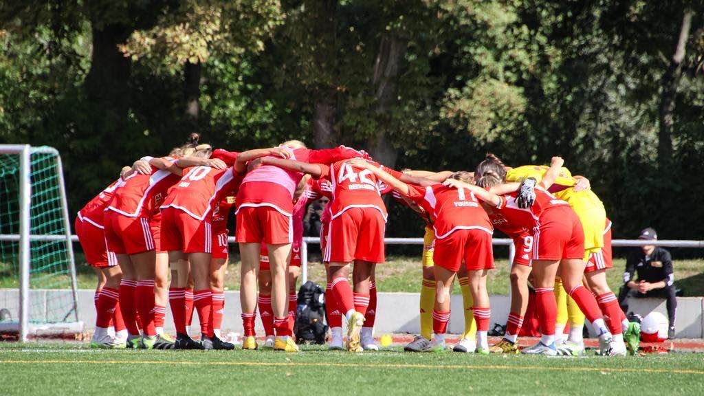 Women's football team in red jerseys forms a circle and discusses before the game on the field.