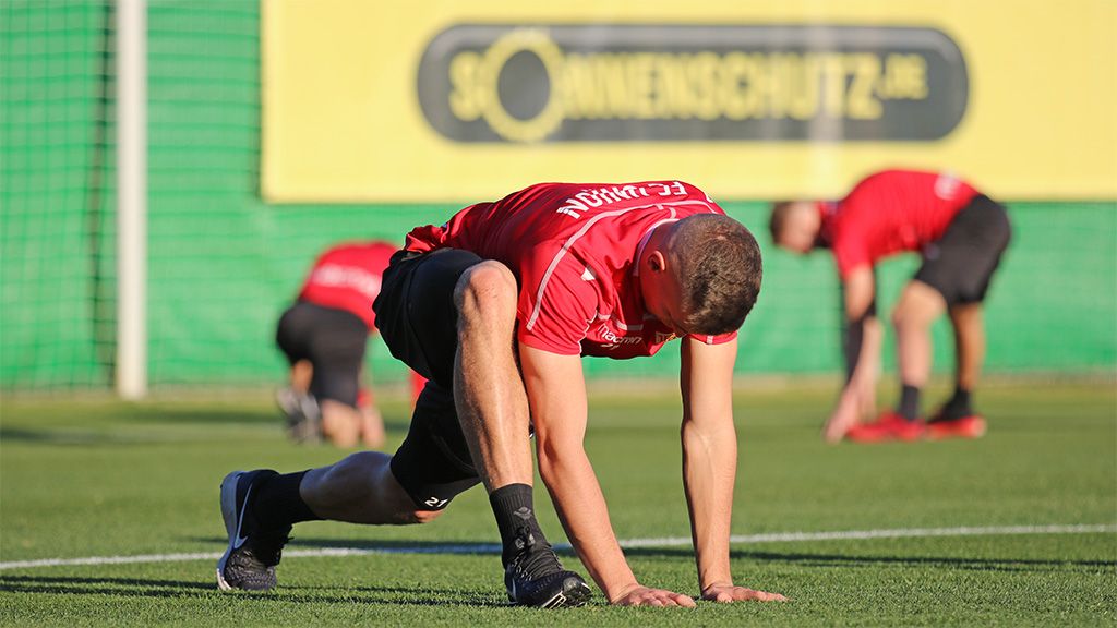 Fußballspieler dehnen sich auf einem Trainingsplatz mit Kunstrasen im Hintergrund.