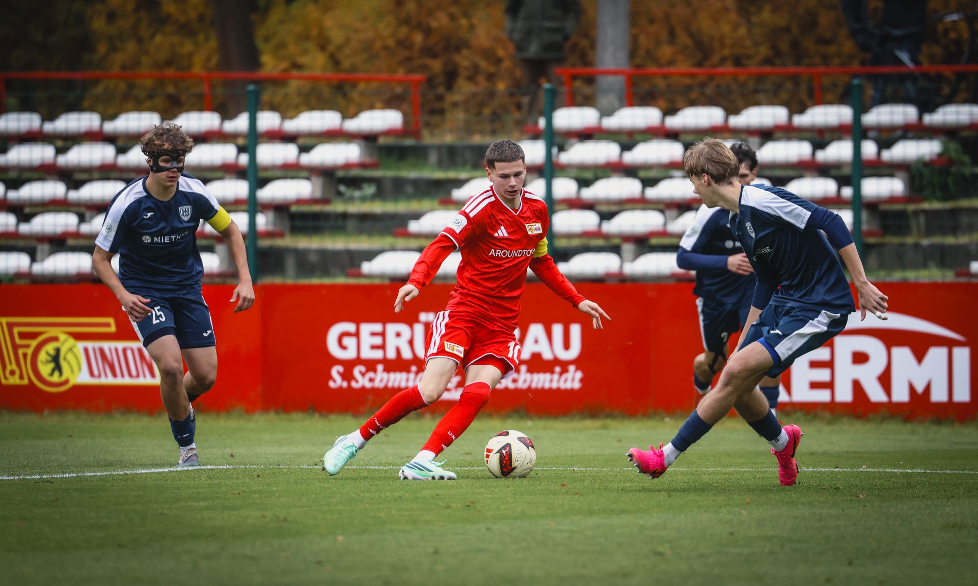 Ein Jugendfußballspiel, bei dem ein Spieler in rotem Trikot den Ball dribbelt, während zwei gegnerische Spieler ihn anvisieren.