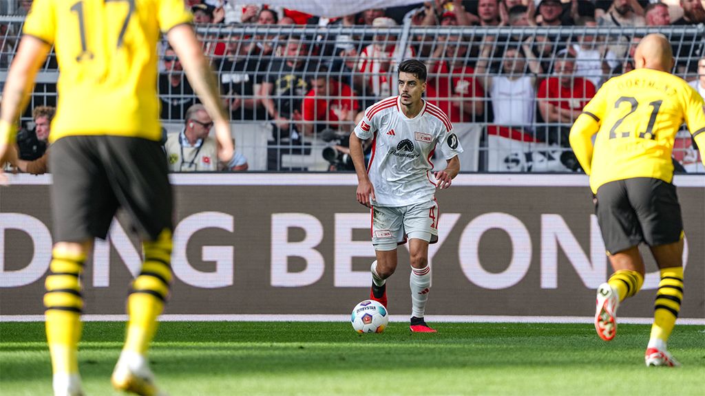 Ein Fußballspieler in weißem Trikot dribbelt den Ball, während zwei Gegner in gelben Trikots ihn beobachten.