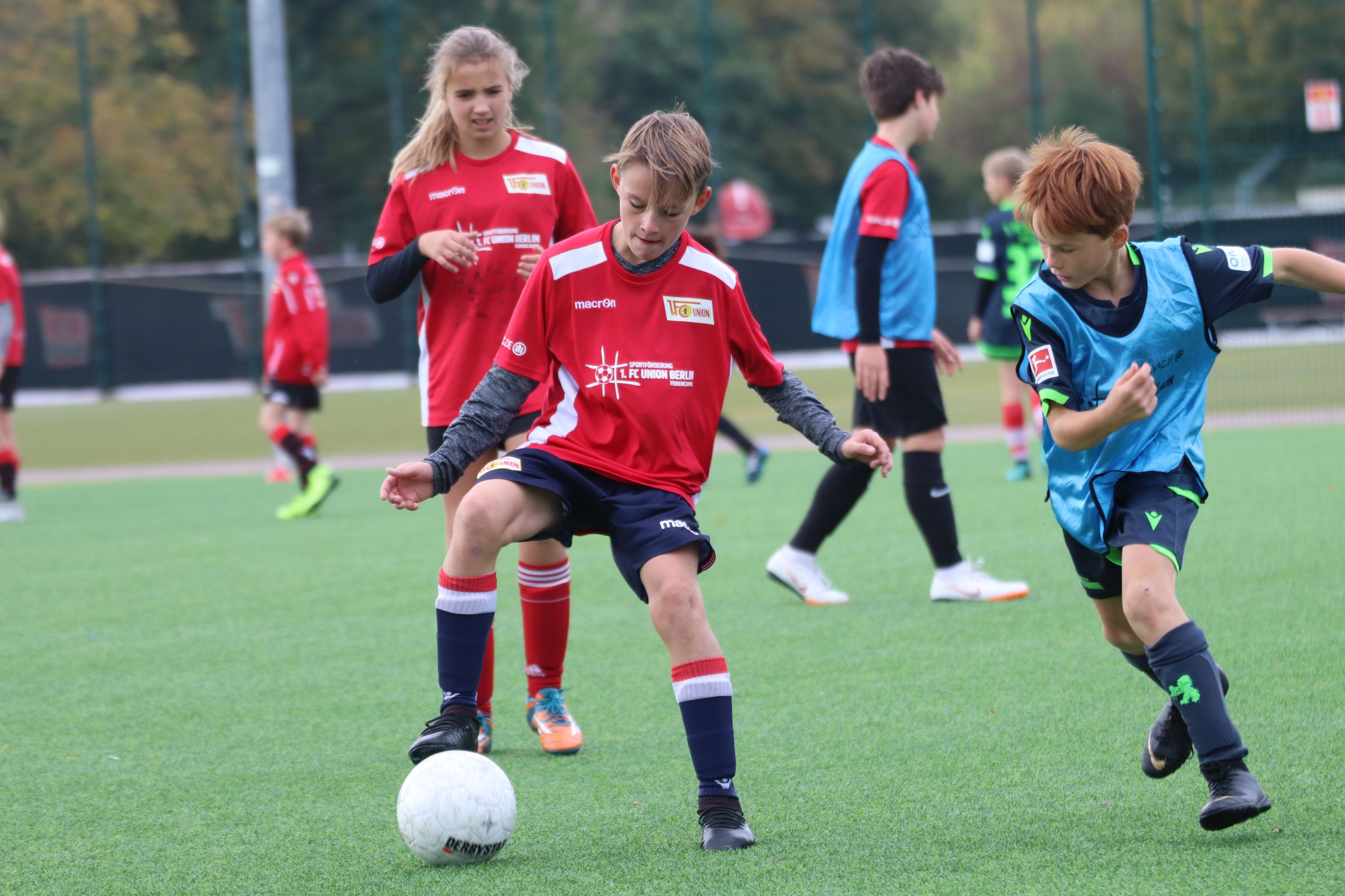 Zwei Kinder spielen Fußball auf einem Kunstrasenplatz, während andere Spieler im Hintergrund aktiv sind.