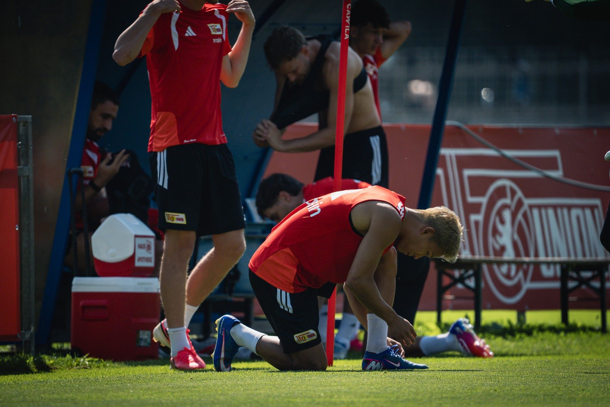 Fußballspieler in Trainingskleidung kniet auf dem Platz und bindet seine Schuhe, während andere Spieler im Hintergrund aktiv sind.