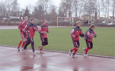 Fünf Sportler in roten Trainingsanzügen joggen auf einem Sportplatz. Im Hintergrund sind Tore und Bäume zu sehen.