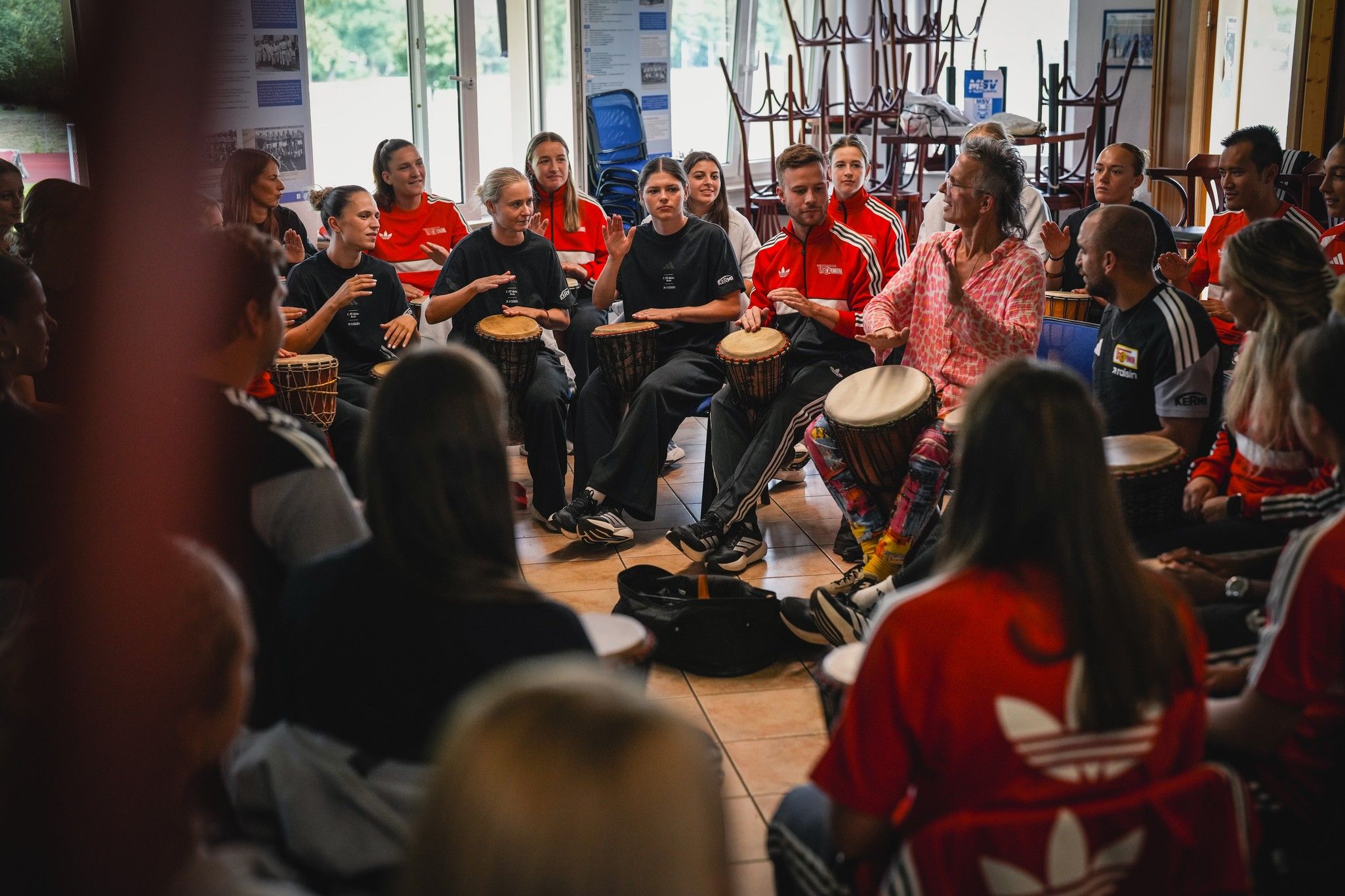 Eine Gruppe von Menschen sitzt in einem Kreis, spielt verschiedene Trommeln und musiziert gemeinsam in einem hellen Raum.