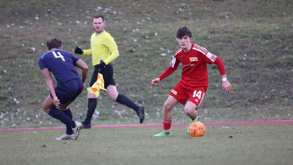 Ein Fußballspieler in roter Uniform dribbelt den Ball, während ein Gegner in Blau ihn verfolgt. Ein Schiedsrichter in Gelb steht im Hintergrund.