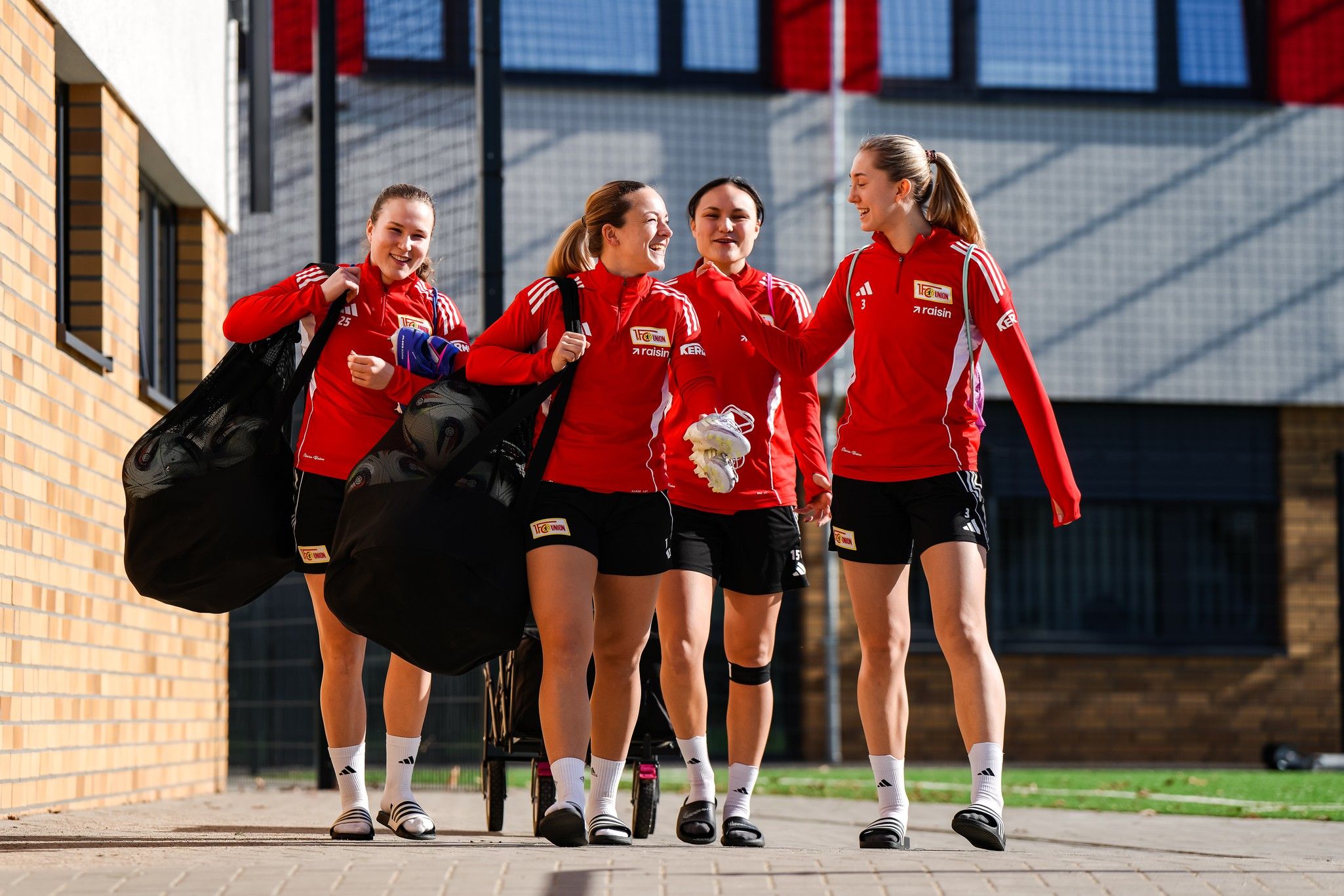Vier Fußballspielerinnen in roten Trainingsjacken tragen Sporttaschen und lachen, während sie sich auf einem Platz unterhalten.