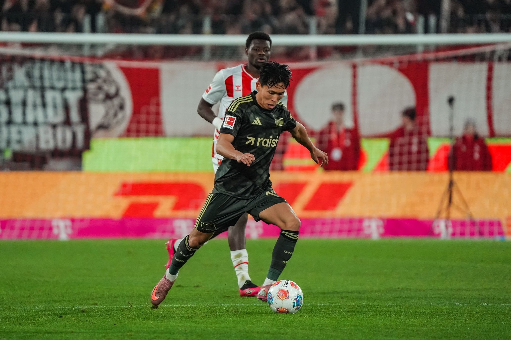 Spielszene im Fußball: Ein Spieler in schwarzem Trikot dribbelt den Ball, während ein gegnerischer Spieler in Weiß verteidigt.