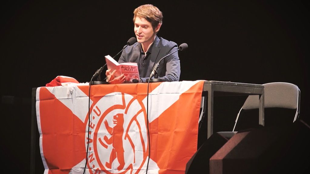 Ein Mann sitzt an einem Tisch mit einer roten und weißen Flagge und liest aus einem Buch.