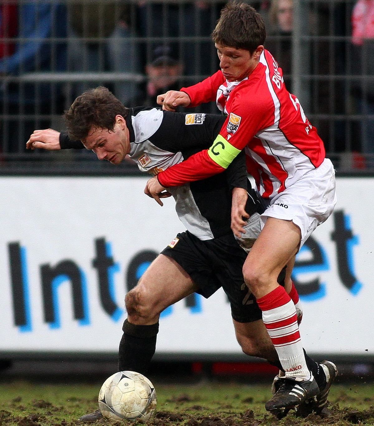 Zwei Fußballspieler kämpfen um den Ball, während einer den anderen von hinten festhält. Beide tragen Trikots in unterschiedlichen Farben.