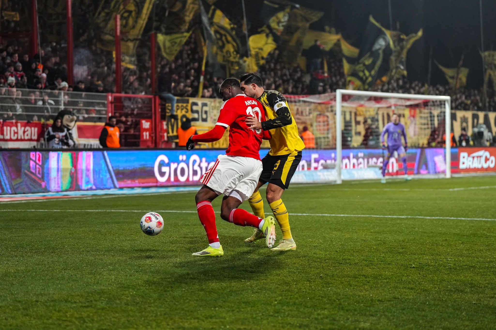Ein Fußballspieler in roter Kleidung dribbelt den Ball, während ein gegnerischer Spieler in gelber Kleidung ihn verteidigt.