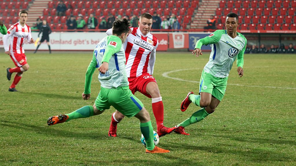 A soccer player in red is dribbling the ball between two players in green jerseys on a field.