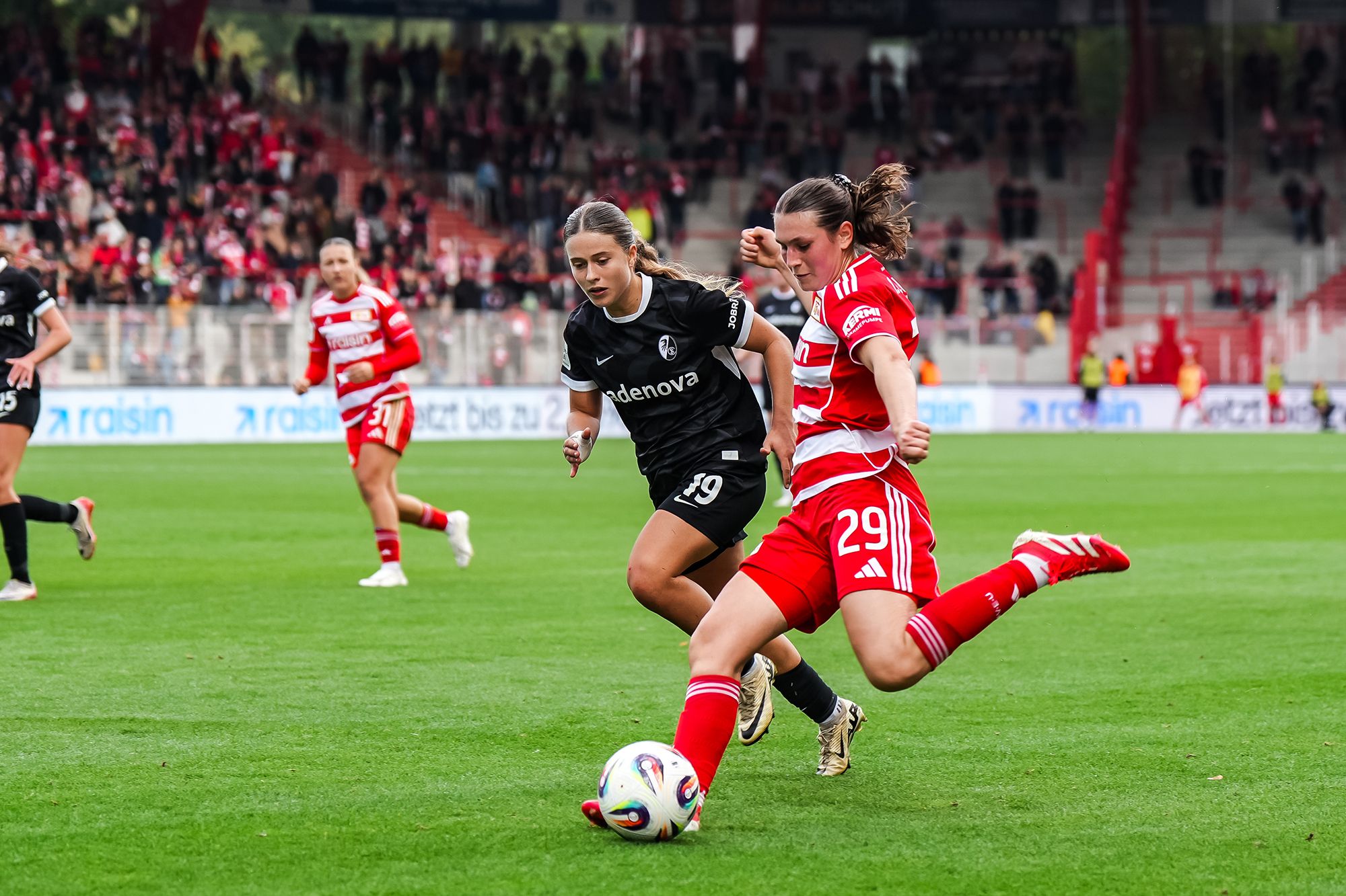 Zwei Fußballspielerinnen in Aktion auf dem Spielfeld, eine in schwarzem Trikot, die andere in rotem Trikot, im Spiel deutliche Bewegung.
