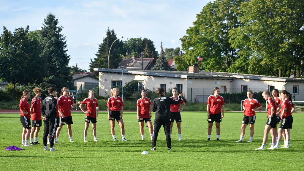 Eine Gruppe von Fußballspielern in roten Trikots versammelt sich auf einem Sportplatz, während ein Trainer Anweisungen gibt.
