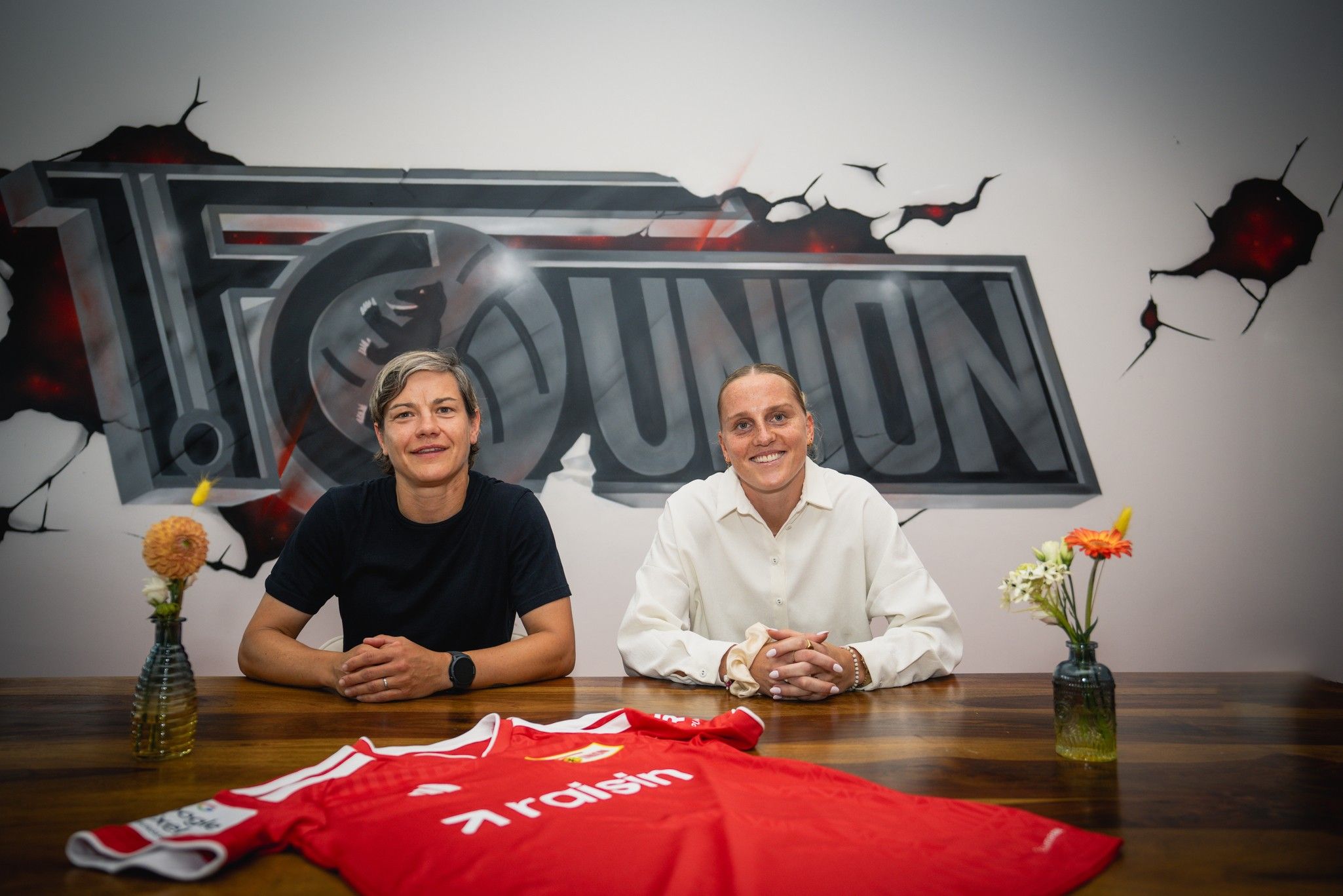 Zwei Frauen sitzen an einem Tisch mit einem roten Fußballtrikot, Blumenvasen und einem Hintergrund mit dem Logo von Union Berlin.