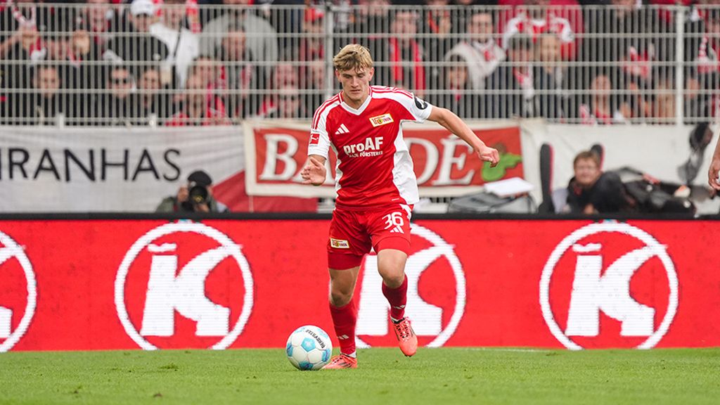 A soccer player in red team gear dribbles the ball on a field surrounded by cheering fans.