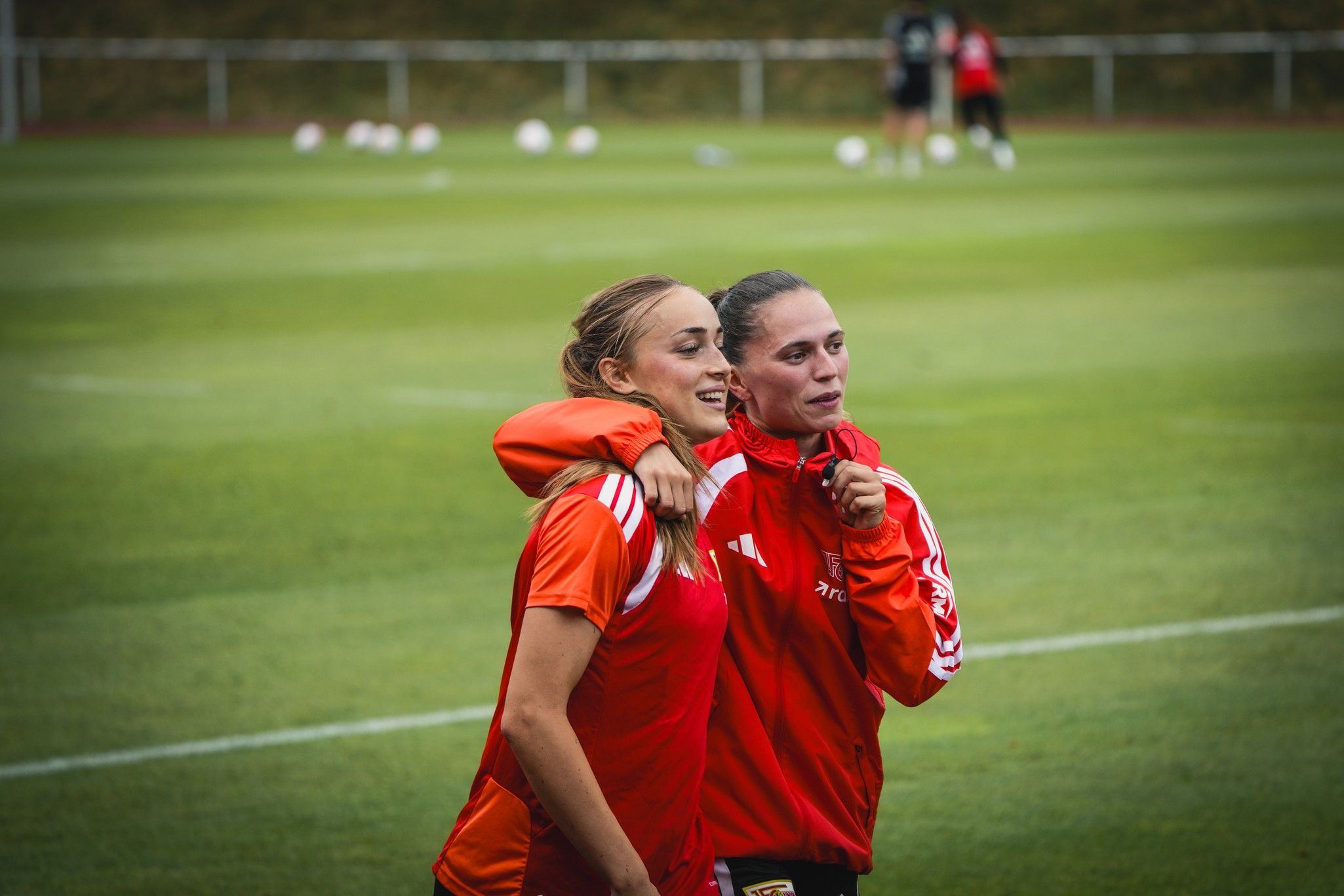 Zwei Frauen in Sportkleidung stehen auf einem Fußballplatz und umarmen sich lächelnd, im Hintergrund weitere Spielerinnen beim Training.
