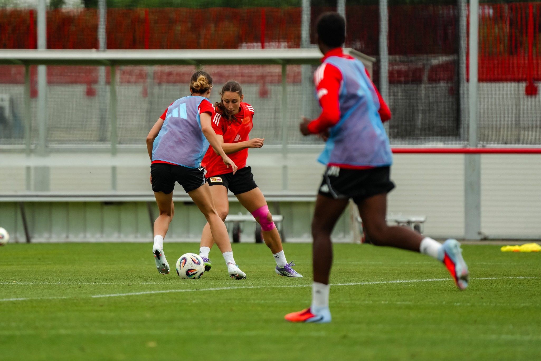 Zwei Frauen im Fußballtrikot spielen auf einem Trainingsplatz, während eine dritte Spielerin im Hintergrund läuft.