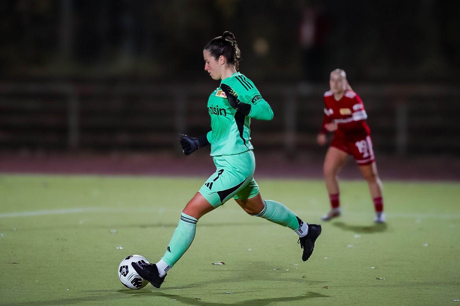Torhüterin in mintgrüner Ausrüstung kickt den Ball, während eine Spielerin in roter Uniform im Hintergrund steht.