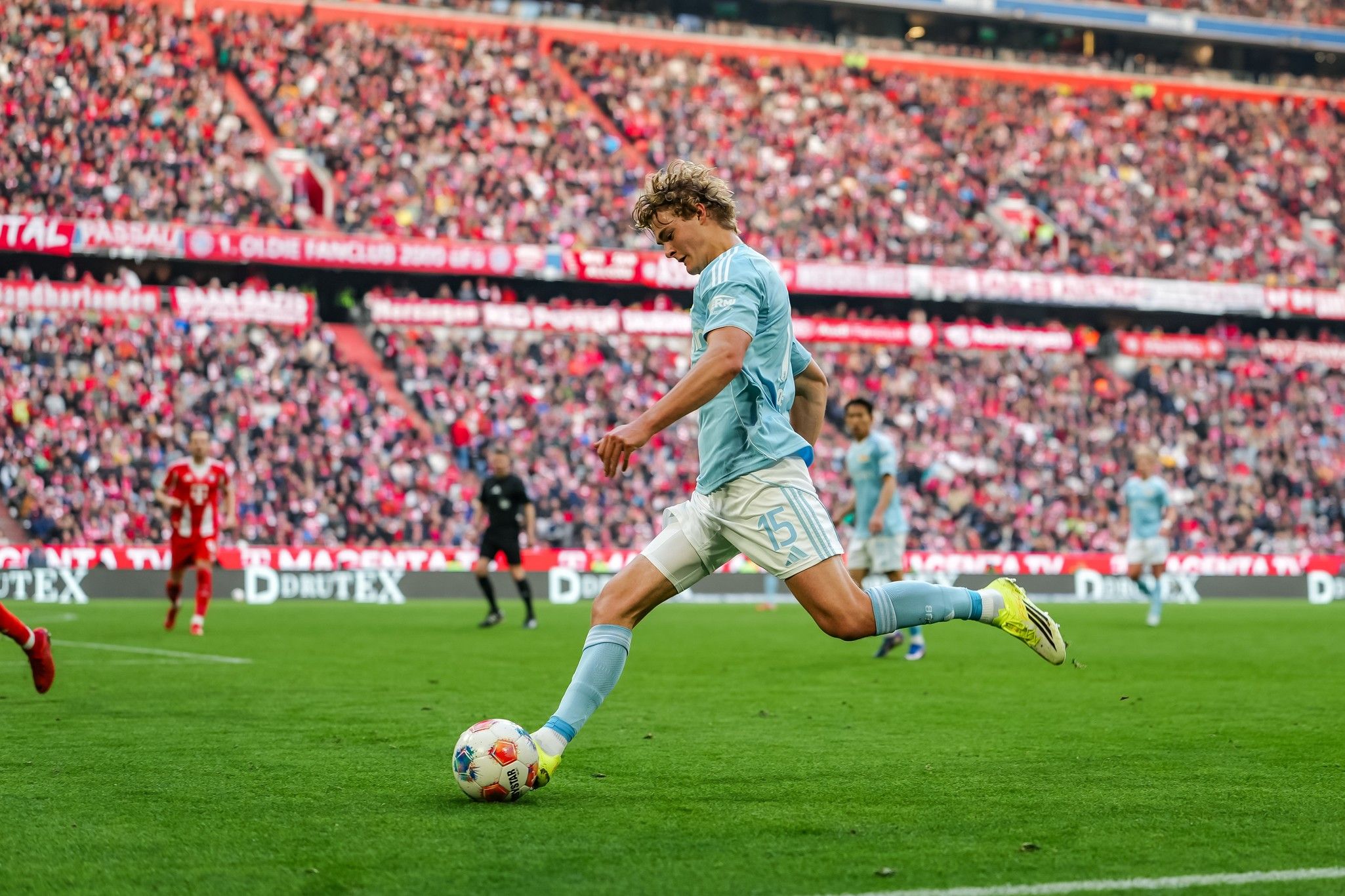 Ein Fußballspieler in hellblauer Ausrüstung tritt den Ball auf einem Rasenplatz, umgeben von jubelnden Fans im Stadion.