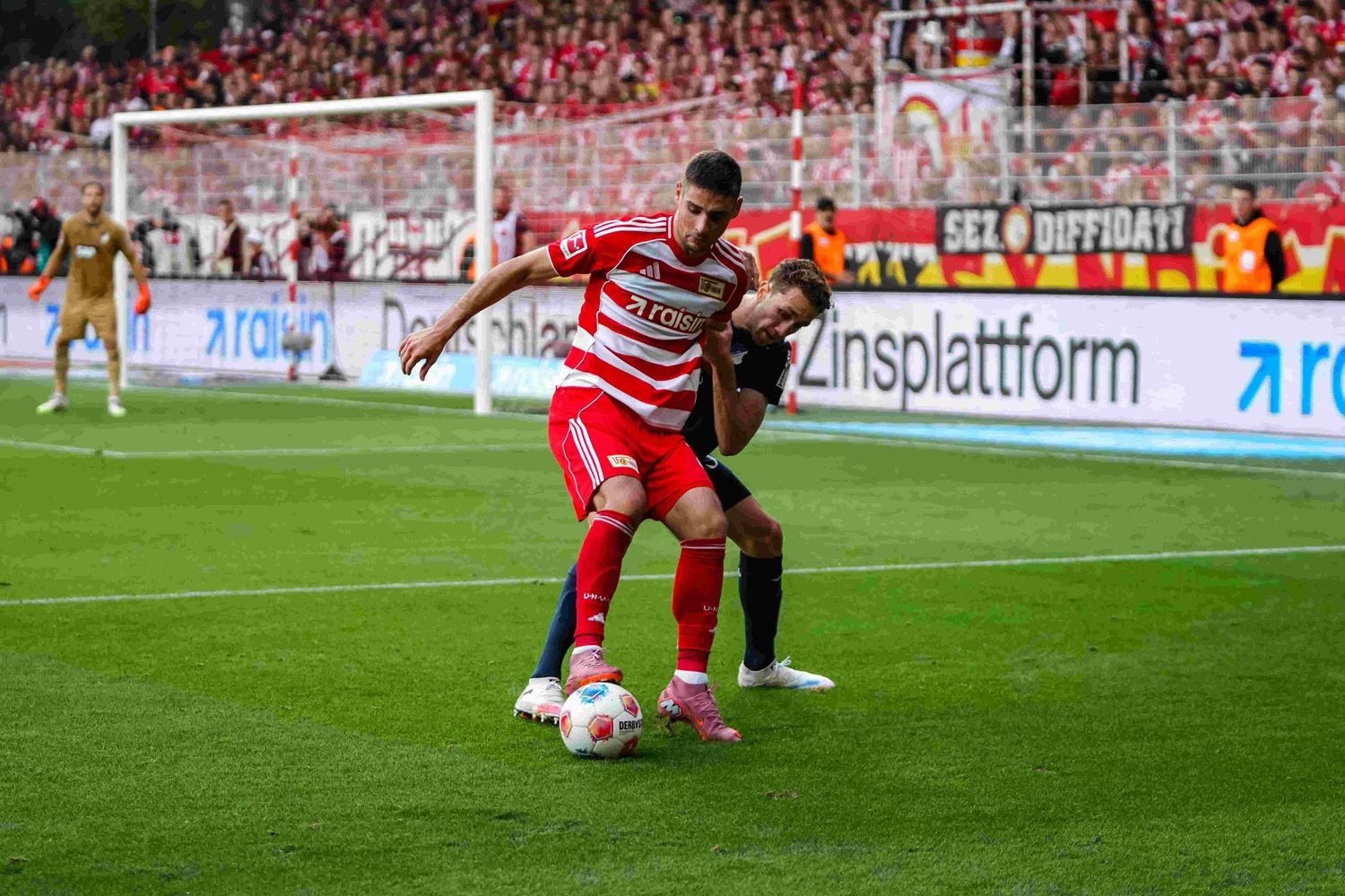 Fußballspieler im roten Trikot kämpft um den Ball, während er von einem Gegner angegrifen wird. Fans sind im Hintergrund sichtbar.