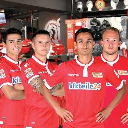 Vier Fußballspieler in roten Trikots, stehen aufrecht und verschränkt die Arme, vor einem Sportartikelgeschäft.