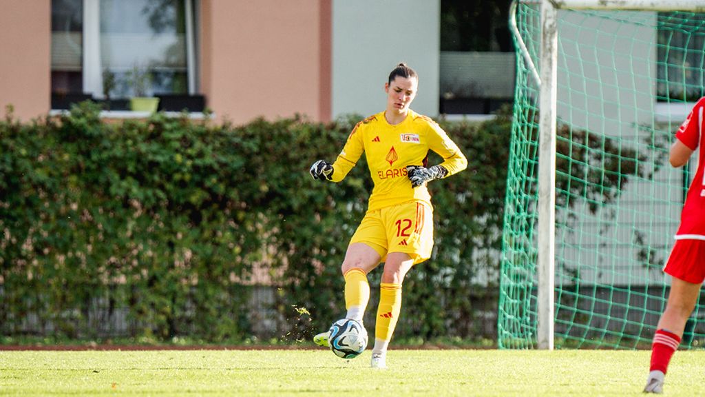 Eine Torhüterin in gelber Uniform steht auf einem Fußballfeld, bereit einen Abschlag zu machen. Im Hintergrund sind Spielerinnen zu sehen.