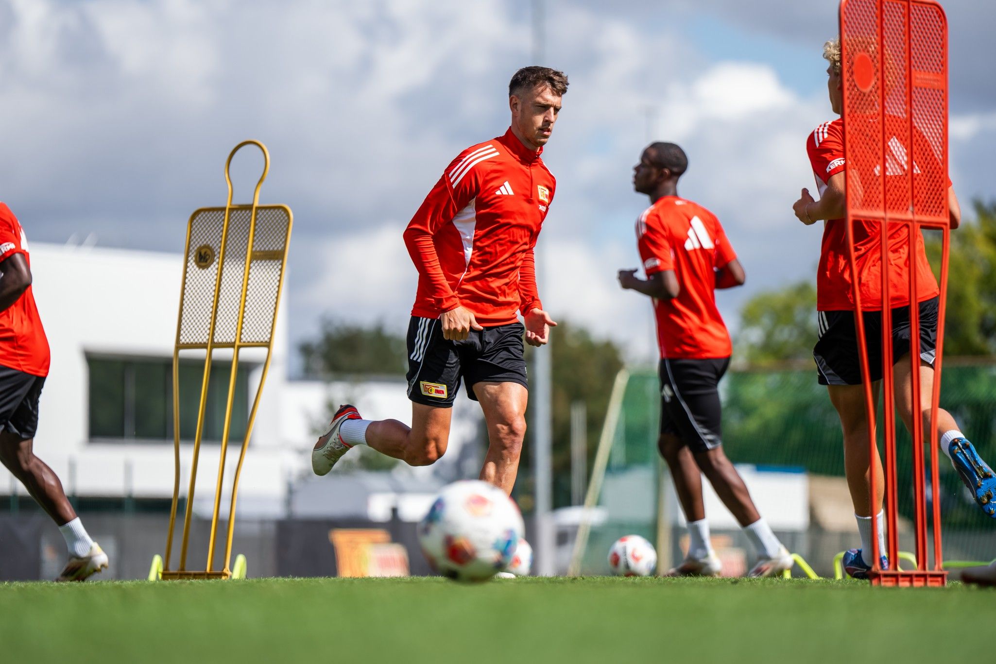 Ein Fußballspieler in rotem Trikot trainiert auf einem Kunstrasenplatz, während andere Spieler im Hintergrund aktiv sind.