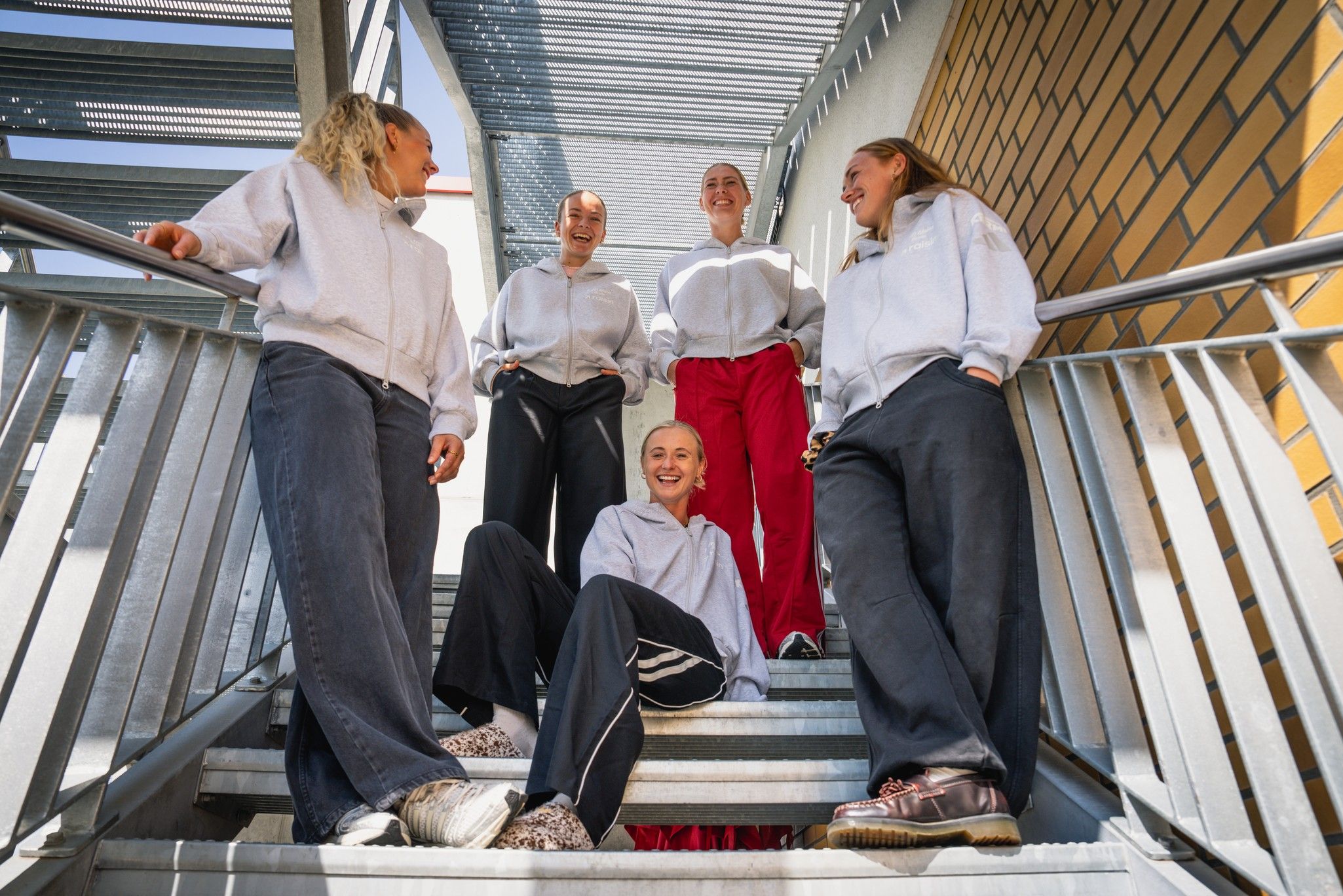 Fünf Frauen sitzen und stehen auf einer Treppe, tragen lässige Kleidung und lachen in Richtung Kamera.