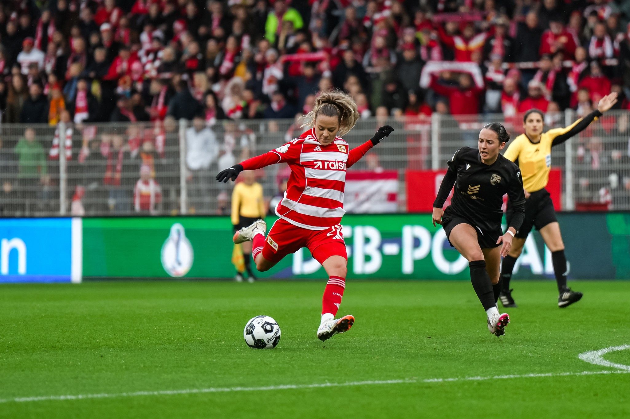 Spielszene im Fußball: Eine Spielerin in rot-weißer Uniform schießt den Ball, während eine schwarz gekleidete Spielerin und eine Schiedsrichterin zuschauen.