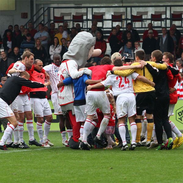 Zwei Fußballmannschaften feiern auf dem Spielfeld zusammen, während eine Maskottchenfigur in der Mitte steht.